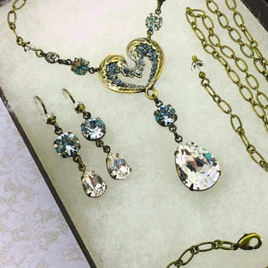 May include: A vintage-style jewellery set comprising a necklace and earrings. The necklace features a heart-shaped pendant with blue accents and a large teardrop crystal. The earrings have matching crystals. The set is gold-toned.