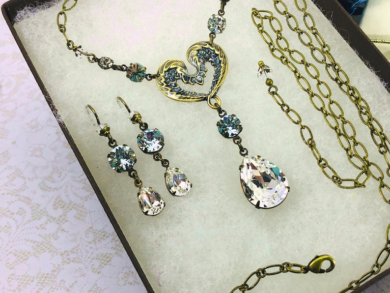 May include: A vintage-style jewellery set comprising a necklace and earrings. The necklace features a heart-shaped pendant with blue accents and a large teardrop crystal. The earrings have matching crystals. The set is gold-toned.