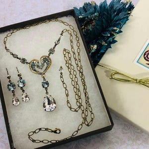 May include: A vintage-style jewellery set in antique gold, comprising a necklace, earrings, and bracelet, displayed in a gift box. The necklace has a heart pendant with blue detailing and teardrop crystals. A gift box with a gold bow is also present.
