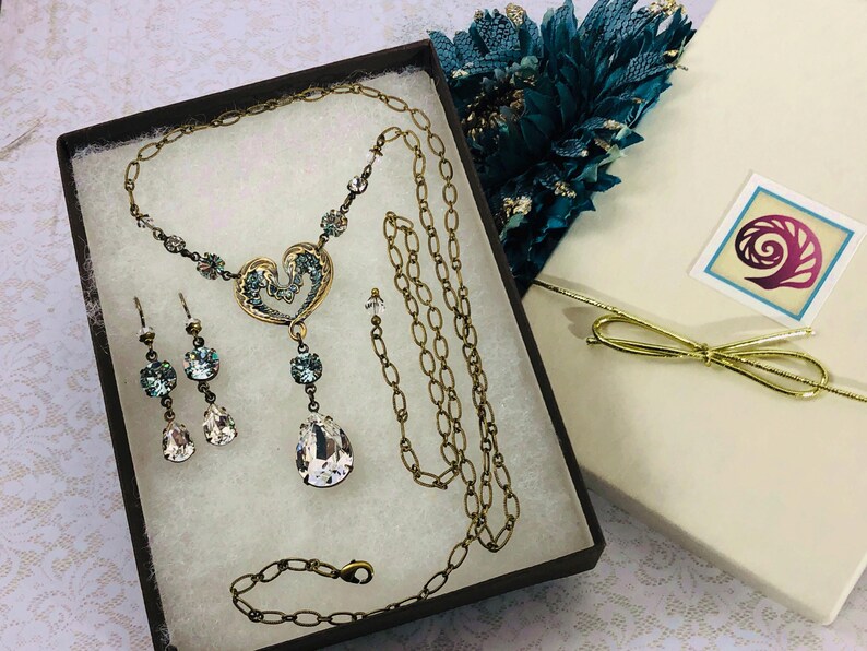 May include: A vintage-style jewellery set in antique gold, comprising a necklace, earrings, and bracelet, displayed in a gift box. The necklace has a heart pendant with blue detailing and teardrop crystals. A gift box with a gold bow is also present.