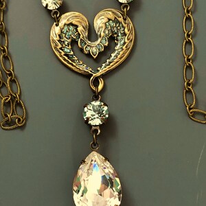 May include: A vintage-style necklace featuring a gold-toned heart pendant with teal accents. A large, clear teardrop crystal hangs below, with smaller crystals along the chain. The necklace has an antique aesthetic.