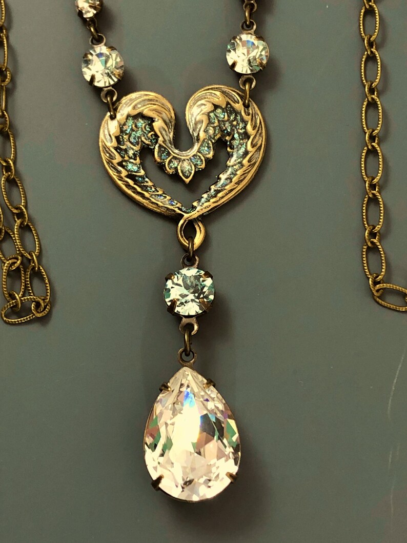 May include: A vintage-style necklace featuring a gold-toned heart pendant with teal accents. A large, clear teardrop crystal hangs below, with smaller crystals along the chain. The necklace has an antique aesthetic.