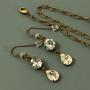 May include: An antique brass-toned jewellery set featuring a necklace and a pair of earrings. The earrings have a round crystal and a teardrop-shaped crystal. The necklace has a chain with small crystals.