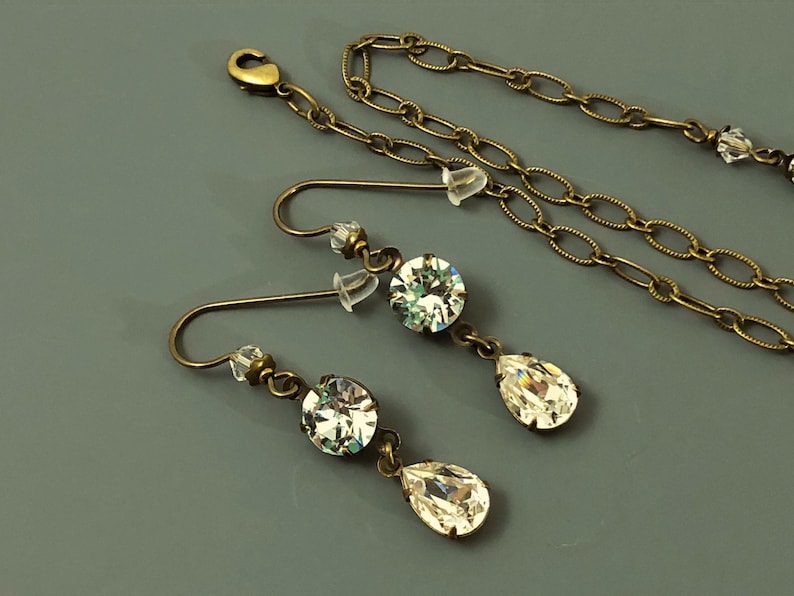 May include: An antique brass-toned jewellery set featuring a necklace and a pair of earrings. The earrings have a round crystal and a teardrop-shaped crystal. The necklace has a chain with small crystals.