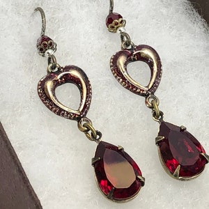 May include: A pair of dangle earrings with a heart-shaped charm and a teardrop-shaped red gemstone. The earrings are made of gold-toned metal and have a vintage style.