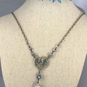 May include: A vintage-style necklace with a heart-shaped pendant and a large, clear teardrop-shaped crystal. The necklace features small, light blue crystals and a delicate chain. The pendant has an ornate design with light blue accents. The necklace is displayed on a mannequin.