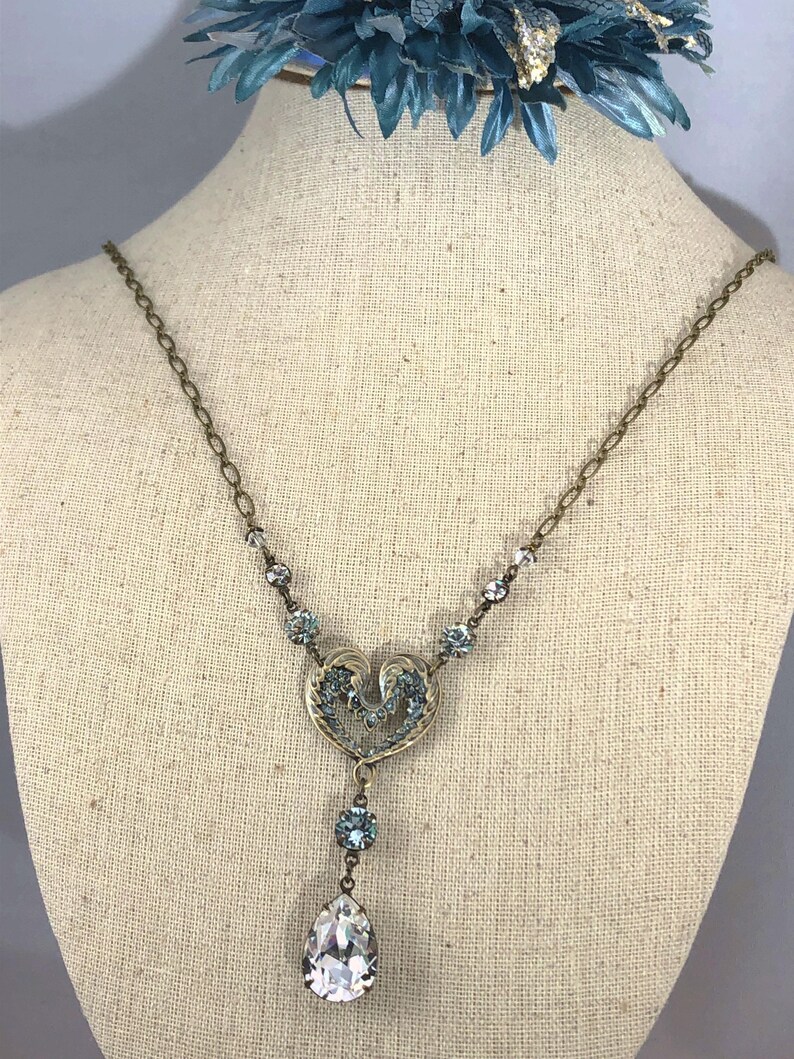 May include: A vintage-style necklace with a heart-shaped pendant and a large, clear teardrop-shaped crystal. The necklace features small, light blue crystals and a delicate chain. The pendant has an ornate design with light blue accents. The necklace is displayed on a mannequin.