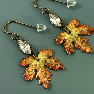 May include: A pair of dangle earrings featuring a leaf design. Each earring has a clear, oval-shaped crystal and a detailed maple leaf pendant in shades of gold, orange, and bronze. The earrings have a hook closure.