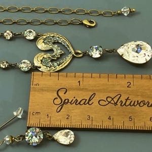 May include: A vintage-style jewellery set featuring a necklace with a heart-shaped pendant and a pair of earrings. The necklace and earrings have clear crystal accents and a teardrop-shaped crystal. The set is displayed with a wooden ruler.