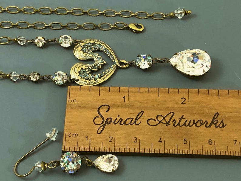 May include: A vintage-style jewellery set featuring a necklace with a heart-shaped pendant and a pair of earrings. The necklace and earrings have clear crystal accents and a teardrop-shaped crystal. The set is displayed with a wooden ruler.