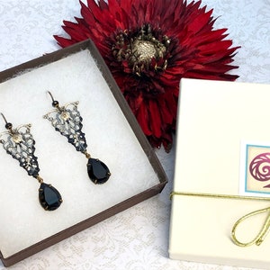 May include: A pair of black and silver filigree earrings with black teardrop-shaped stones. The earrings are in a small brown box with white padding. The box is tied with a gold ribbon.