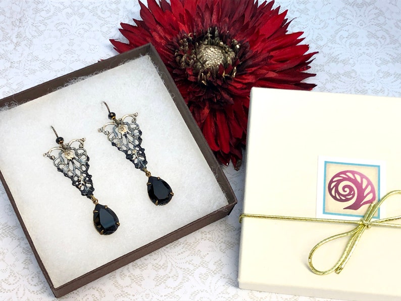 May include: A pair of black and silver filigree earrings with black teardrop-shaped stones. The earrings are in a small brown box with white padding. The box is tied with a gold ribbon.
