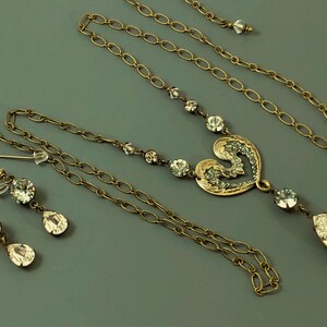 May include: A vintage jewellery set comprising a necklace and earrings. The necklace features a heart-shaped pendant with clear crystals and a teardrop crystal. The earrings have round and teardrop crystals. The set is made of gold-toned metal.
