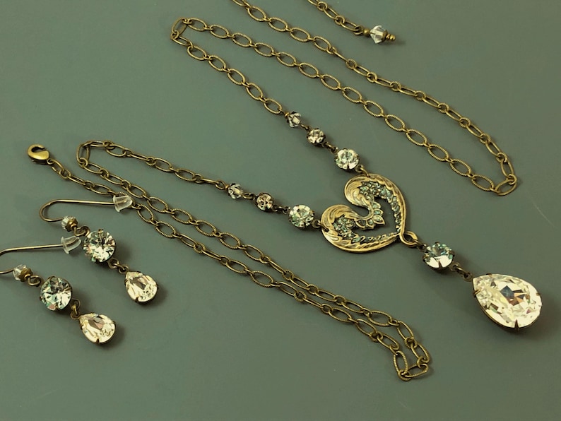 May include: A vintage jewellery set comprising a necklace and earrings. The necklace features a heart-shaped pendant with clear crystals and a teardrop crystal. The earrings have round and teardrop crystals. The set is made of gold-toned metal.