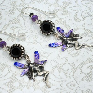May include: A pair of silver-toned fairy earrings. Each earring features a black, faceted bead with a decorative silver setting, purple and black beads, and a fairy charm with blue and purple wings. The earrings have a hook closure.
