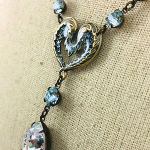 May include: A vintage necklace with a heart-shaped pendant in blue and gold tones. It features a teardrop crystal and smaller, round crystals on the chain. The metal has an antique finish, creating a classic look.