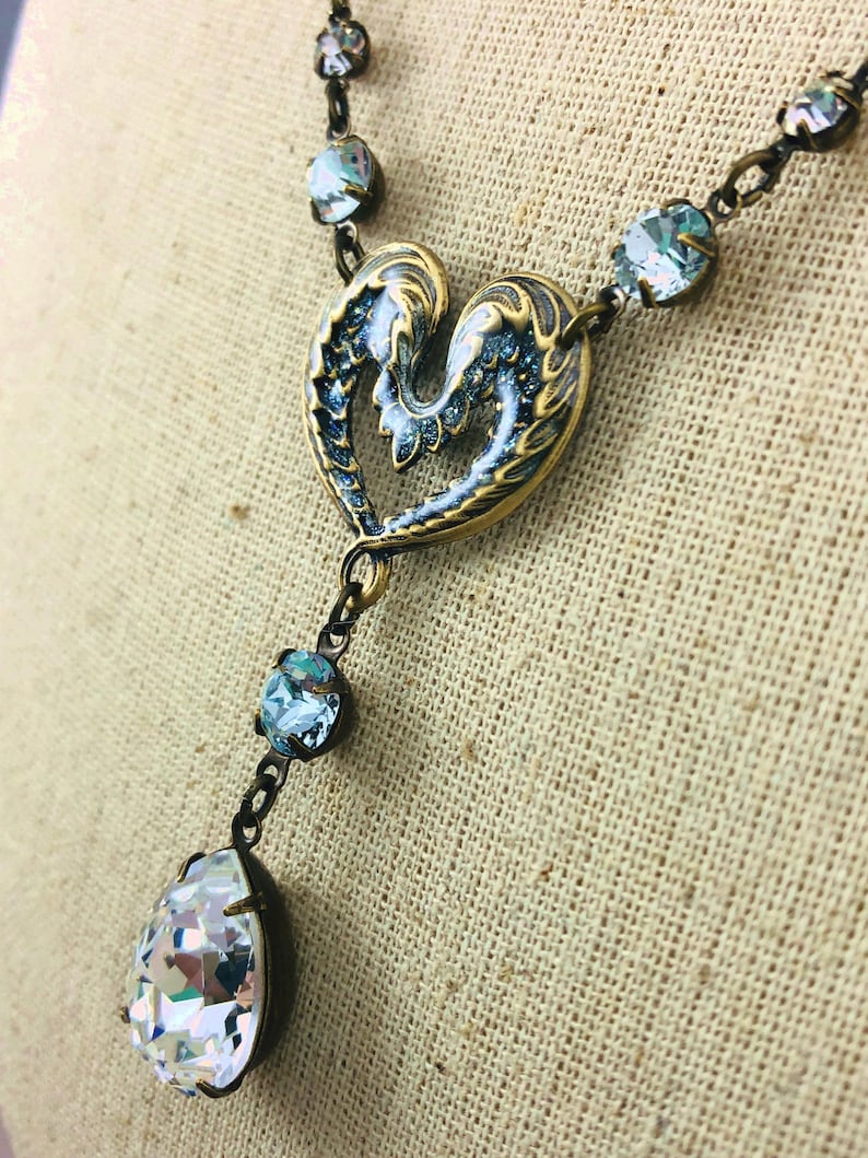 May include: A vintage necklace with a heart-shaped pendant in blue and gold tones. It features a teardrop crystal and smaller, round crystals on the chain. The metal has an antique finish, creating a classic look.