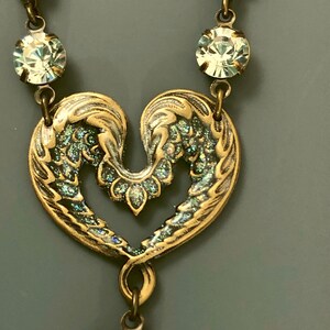 May include: A vintage necklace with a heart-shaped pendant, detailed with wing-like designs and a bronze finish with iridescent highlights. The necklace includes clear, round crystals along the chain and at the pendant's base, adding a touch of sparkle.