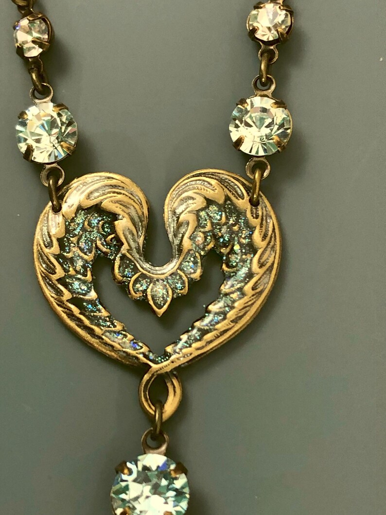May include: A vintage necklace with a heart-shaped pendant, detailed with wing-like designs and a bronze finish with iridescent highlights. The necklace includes clear, round crystals along the chain and at the pendant's base, adding a touch of sparkle.