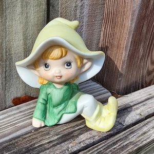May include: A ceramic figurine of a seated elf with a yellow hat and shoes, and a green tunic. The elf has blonde hair and is resting on a weathered wooden surface. The figurine has a whimsical, fairytale aesthetic.