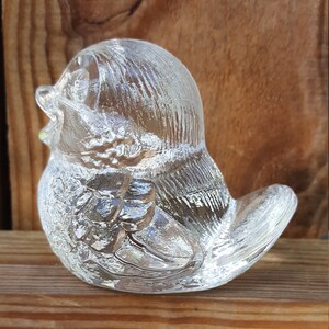 May include: A clear glass bird figurine with textured details. The bird is in a sitting position, with its beak open. The figurine is displayed on a wooden surface, showcasing its transparent design.