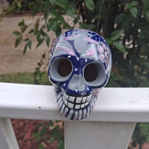 May include: A decorative ceramic skull with a blue, gray, and pink floral design. The skull has large eye sockets and a detailed, smiling mouth. The skull is resting on a white surface.