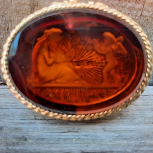 May include: Oval pendant with a detailed amber-colored cameo depicting classical figures. The pendant is set in a gold-tone frame with a twisted rope design. The cameo features a woman and a cherub with a sunburst background.