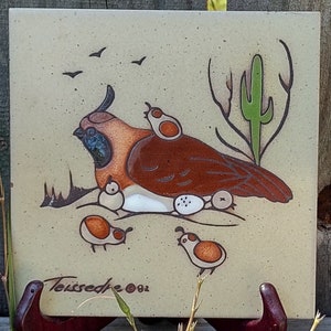 May include: A hand-painted ceramic tile featuring a brown and orange quail with its chicks. The tile is set against a beige background with a green cactus and a brown branch. The tile is signed "Toureed 2001" in the bottom left corner.