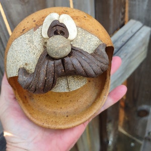 May include: A ceramic dish shaped like a face with a brown mustache, a brown nose, and white eyes. The dish is brown and tan with a speckled glaze.