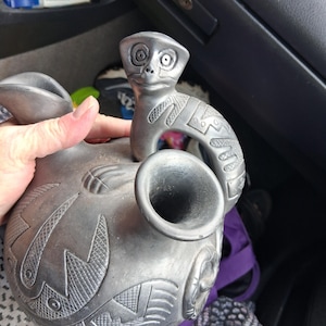May include: A dark gray ceramic vessel featuring a rounded body with etched geometric patterns and a sculpted animal figure attached to the top. The vessel includes a circular opening and a curved handle that connects to the animal's body.