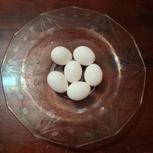 May include: A clear, octagonal glass serving dish with etched floral designs holds six white eggs. The dish sits on a dark wooden surface, creating a contrast between the clear glass and the eggs.