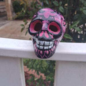 May include: A decorative skull figurine with a black base and pink floral designs. The skull has pink eye sockets and a pink mouth. The teeth are white. The figurine is sitting on a white railing.