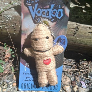 May include: A tan twine-wrapped doll keychain with a small red fabric heart on its chest, black bead eyes, and a thin black neck cord. The doll is attached to a silver ball chain and mounted on a blue and white cardboard backing with the text Voodoo.