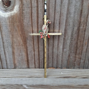 May include: A gold-colored metal cross with a decorative top piece featuring a red and green floral design. The cross is set against a weathered wooden background, with the vertical beam having a textured pattern.