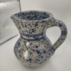 May include: A small, blue and white speckled ceramic pitcher with a handle. The pitcher has a blue stripe around the middle.