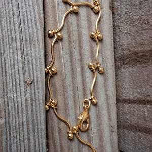 May include: A gold-tone bracelet featuring a delicate, wavy wire chain accented with small, evenly spaced spherical beads. The piece includes a lobster claw clasp and a small, oval-shaped metal tag engraved with the brand name Avon.