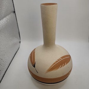 May include: A small, white ceramic vase with a brown and black design of feathers. The vase has a round body and a narrow neck.