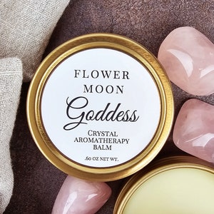 May include: A gold tin with a white label that reads "Flower Moon Goddess Crystal Aromatherapy Balm .60 oz Net Wt.". The tin is surrounded by pink rose quartz crystals.