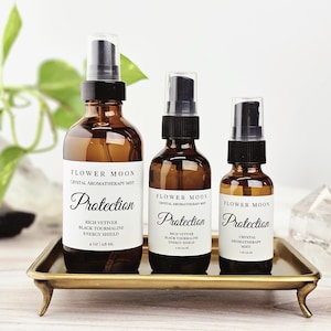 May include: Three amber glass aromatherapy mist bottles with black spray tops are displayed on a gold tray. Each bottle has a white label with the words "Flower Moon" and "Protection". The largest bottle is 4 oz.