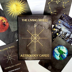 May include: A set of black and gold astrology cards with a black box that says "The Living Wheel Astrology Cards". The cards feature watercolor illustrations of astrological signs and symbols.