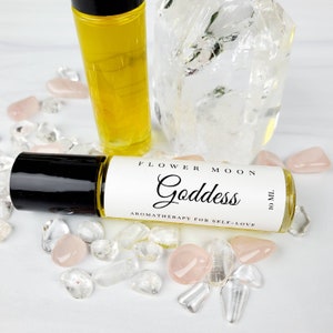 May include: A clear glass roller bottle with a black cap and a white label that reads "FLOWER MOON Goddess AROMATHERAPY FOR SELF-LOVE 10 ML". The bottle is surrounded by clear and pink crystals.