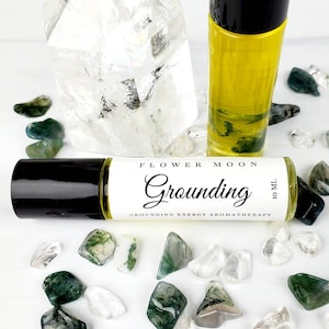 May include: A 10 ml roll-on bottle of grounding essential oil with a white label that says "Flower Moon Grounding Grounding Energy Aromatherapy". The bottle is surrounded by green and clear stones.