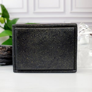 May include: A rectangular black box with a speckled gold pattern. The box has a lid and is made of a solid, possibly stone-like material. The box is sitting on a white surface.