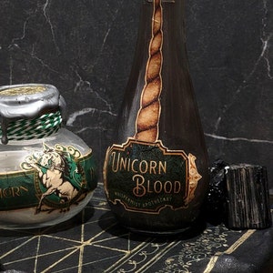 Könnte beinhalten: Zwei Glasflaschen mit silbernen und grünen Wachssiegeln. Die Flaschen enthalten weiße und dunkle Flüssigkeiten. Die Flaschen sind mit "Einhornblut" und "Einhorn" beschriftet.