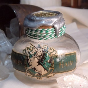 Könnte beinhalten: Eine durchsichtige Glasflasche mit einem grün-weißen Etikett, auf dem "Unicorn Hair" steht und eine Einhorn-Illustration abgebildet ist. Die Flasche ist mit einem silbernen Wachssiegel versehen und um den Hals ist ein grün-weißes Band gebunden.