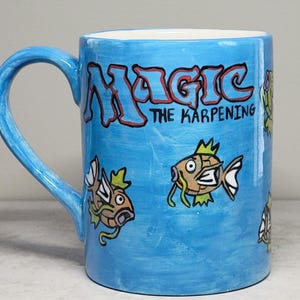 May include: A light blue ceramic mug featuring hand-painted illustrations of orange fish with yellow crowns and green whiskers. The text "MAGIC THE KARPENING" is written in a bold, red-outlined font across the upper portion of the mug.