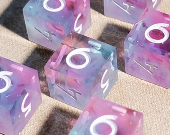 May include: Six translucent dice with a colorful blend of purple, blue, and pink hues. Each die features white numerals and symbols, with the number 6 prominently displayed on top. The dice are arranged on a neutral-toned surface.