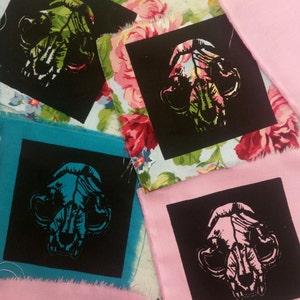 May include: Four fabric squares with black fabric squares on top. Each black square has a white silhouette of an animal skull printed on it. The skulls are in different colors: green, blue, and pink.