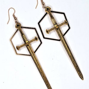 May include: A pair of gold-toned earrings featuring a sword design within a hexagonal frame. The swords are detailed with a textured surface and a pointed tip.