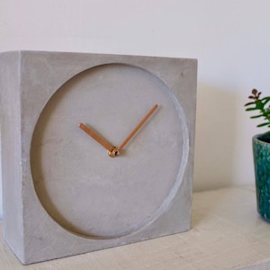 May include: A square concrete clock with a circular face and copper-coloured hands. The clock is light grey and sits on a white surface. A small green potted plant is to the right of the clock.